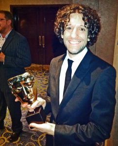 A photo of Greg in a suit, posing with a gold BAFTA award